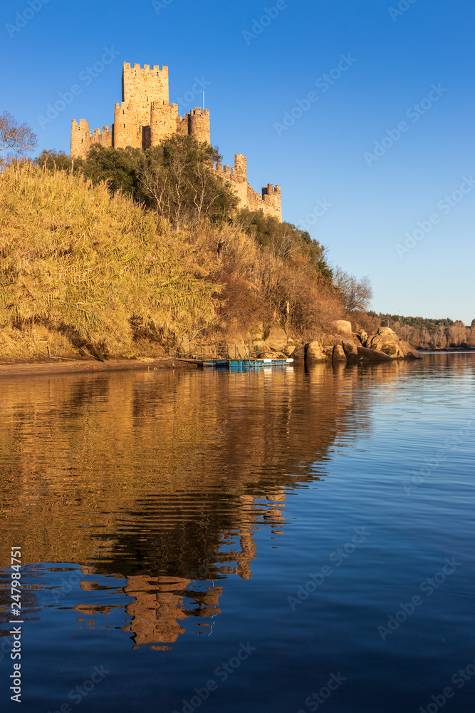 Almourol, Portugal - January 12, 2019: View of the Almourol castle from the Tagus river with the reflection of it in the water, lit by the late afternoon sun with blue sky.