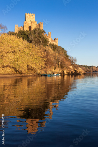Almourol, Portugal - January 12, 2019: View of the Almourol castle from the Tagus river with the reflection of it in the water, lit by the late afternoon sun with blue sky.