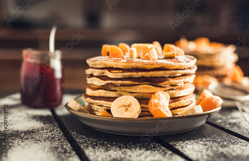 Stack of golden pancakes with bananas and oranges on wooden board covered with caster sugar. Heap of american pancakes with maple syrup and a glass of jam in kitchen.