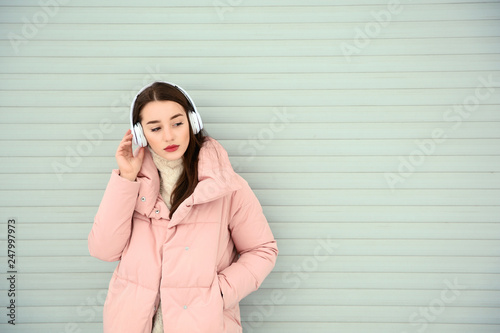 Beautiful young woman listening to music with headphones against light wall. Space for text