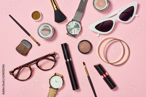 Top view of watches, lipstick, glasses, sunglasses, eye shadow, blush, cosmetic brushes, bracelets and mascara on pink background