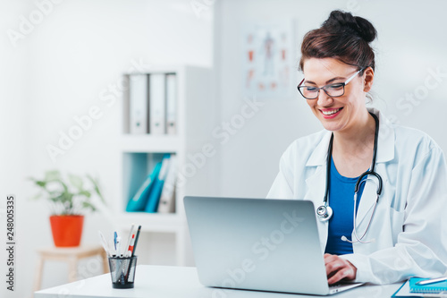 Specialist looking at results from exam on laptop. Friendly woman doctor working on computer in her office