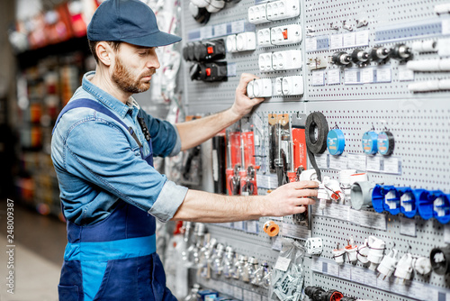 Cuadro en lienzo Handsome electrician in workwear choosing electrical goods in the building shop
