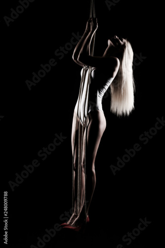 Beautiful slender blonde girl, wearing a white bodisuit and red stilettos, sensually hangs and dances on the ropes. Black background. Artistic noir silhouette photo