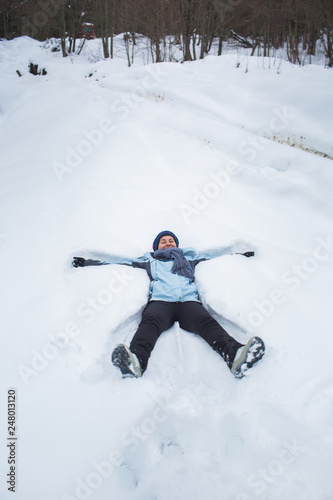 girl in pants, jacket, gloves and scarf lay in the snow in front of the forest macking snow angel