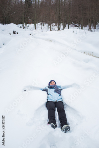 girl in pants, jacket, gloves and scarf lay in the snow in front of the forest macking snow angel