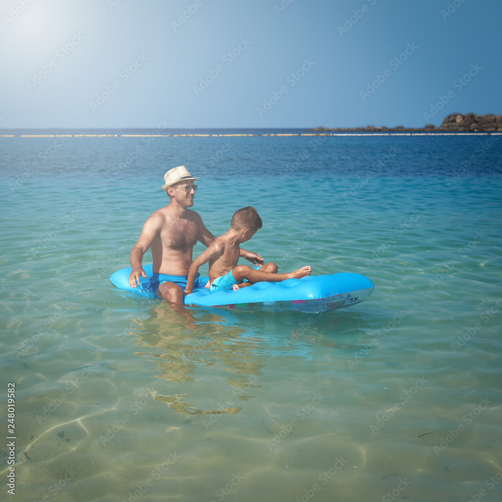 Father and son are swimming on the blue air mattress in the crystal ocean water. They are enjoying their summer holidays.