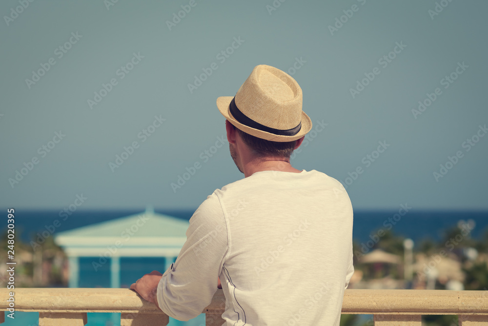 Young European man in sunhat is enjoying wonderful view from the hotel’s balcony.