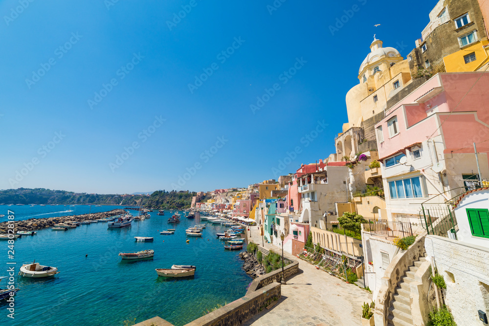 Wide angle photo of vibrantly colorful housing, fishing and sightseeing boats, cafes and restaurants at Marina Corricella in sunny summer weather in Procida island, Italy.