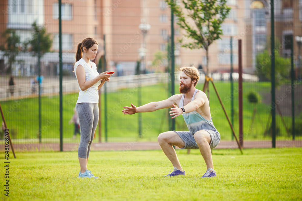 Team work in sports. A female trainer is training a young handsome man with a beard in a park on green grass, a lawn at sunset. A girl holds a phone in her hand and uses a stopwatch