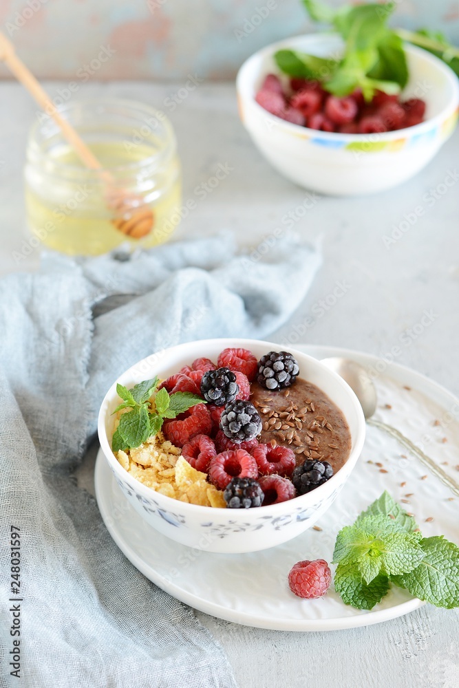 Sunday Breakfast. Oatmeal porridge with banana and flax seed, raspberries, blackberries, corn flakes, honey and mint, selective focus. Useful food, diet. Plate or cup with vegan porridge, smoothies Sunday Breakfast. Oatmeal porridge with banana and flax seed, raspberries, blackberries, corn flakes, honey and mint, selective focus. Useful food, diet. Plate or cup with vegan porridge, smoothies