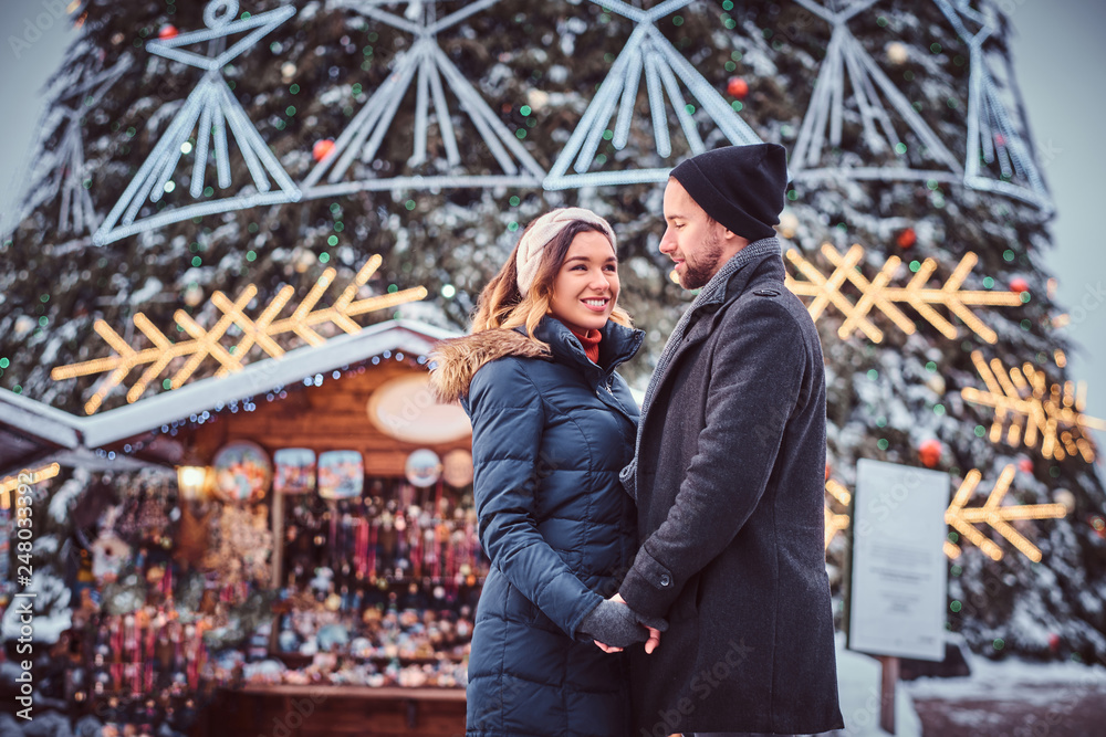 A happy young couple hold hands, enjoying spending time together, standing near a city Christmas tree