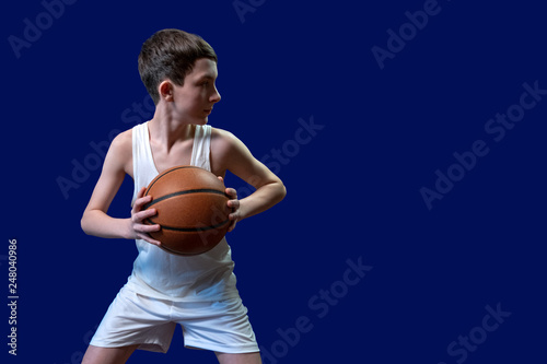 A teenager in a white t-shirt and sports shorts holds a basketball and looks away. Isolated on blue background. Copy space. Blank for sports poster.