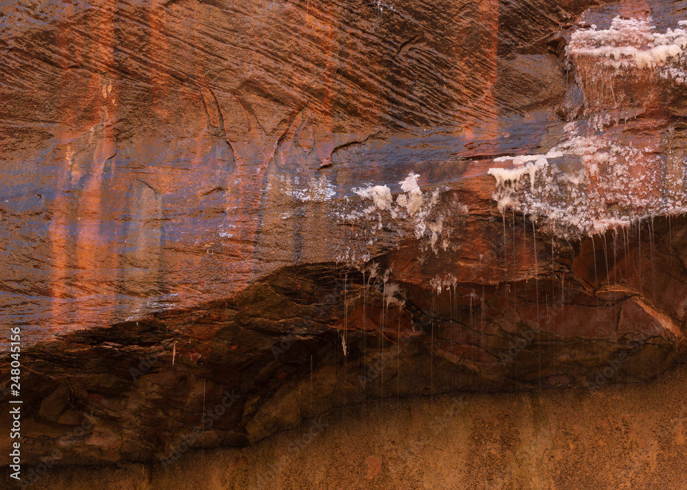 Melting snow drips from the edge of a small blind arch of sandstone streaked with red orange and black.