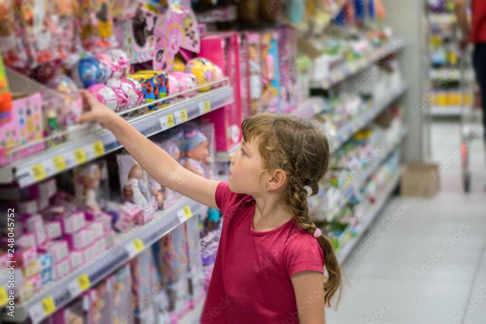 A little girl is buying presents. The child chooses toys in the supermarket. Baby takes the toy from the store shelf.