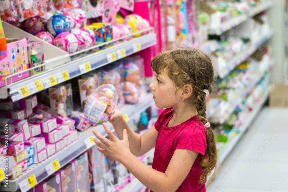 Little girl buys a plastic toy egg in a supermarket. Baby takes a gift from the store shelf. A little girl takes a plastic toy egg in a supermarket Little girl buys a plastic toy egg in a supermarket. Baby takes a gift from the store shelf. A little girl takes a plastic toy egg in a supermarket