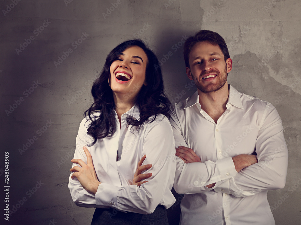 Happy toothy laughing business woman and smiling businessman in white shirts standing with folded arms on loft style wall studio background Happy toothy laughing business woman and smiling businessman in white shirts standing with folded arms on loft style wall studio background