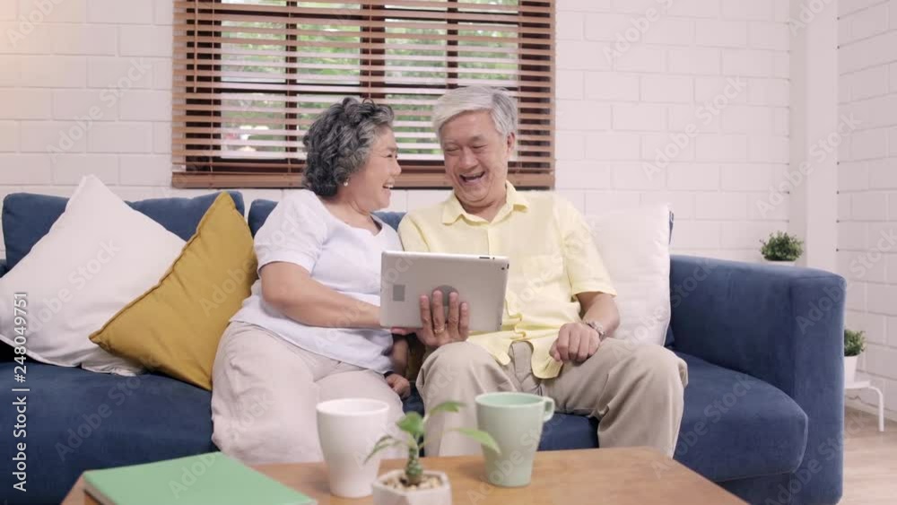 Asian elderly couple using tablet watching TV in living room at home, couple enjoy love moment while lying on sofa when relaxed at home. Enjoying time lifestyle senior family at home concept. Asian elderly couple using tablet watching TV in living room at home, couple enjoy love moment while lying on sofa when relaxed at home. Enjoying time lifestyle senior family at home concept.