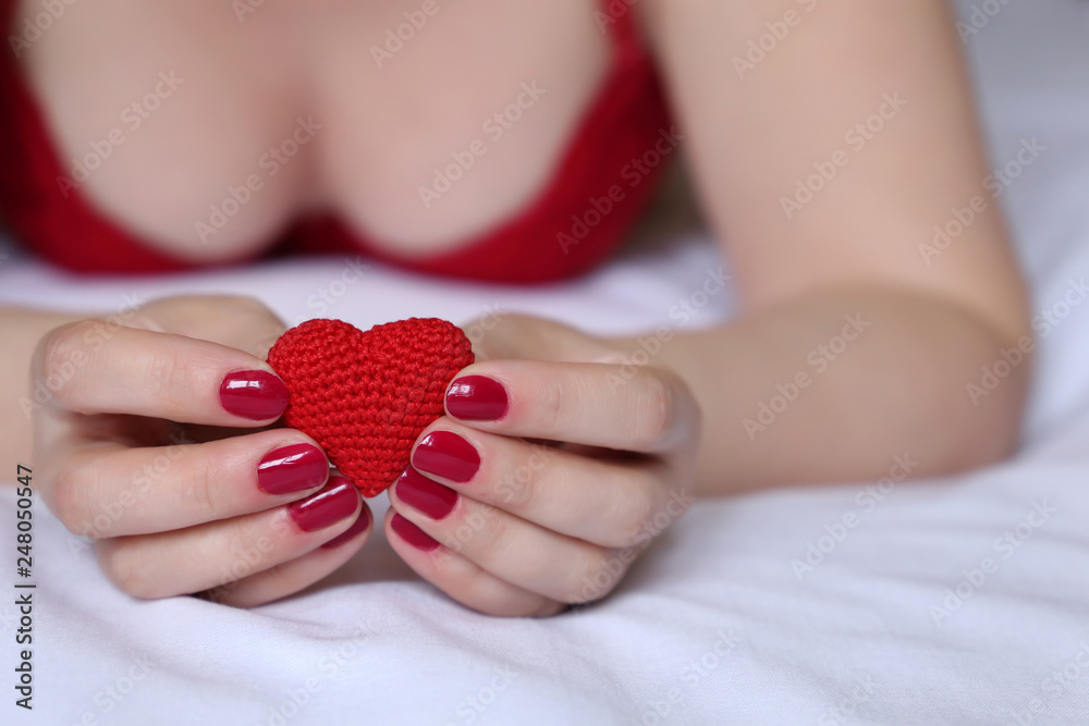 Valentine heart in female hands, declaration of love. Sexy woman in red bra  lying on the bed with knitted symbol of love, concept of romantic sex,  celebration, health care Stock Photo
