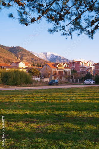 Beautiful winter Mediterranean landscape with a village, snow-capped mountains and long shadows from trees on green grass. Montenegro, Tivat, the village of Donja Lastva