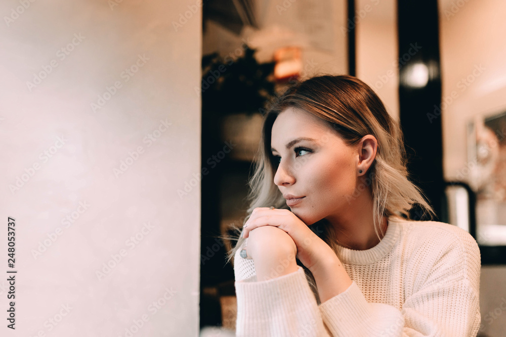 Fototapeta premium Cute blonde girl sits in a cafe looking out the window and romantically meets the morning.