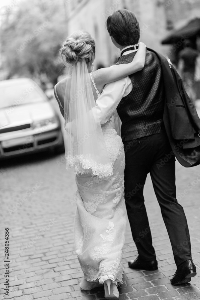 stylish bride and groom walking in city street. happy luxury wedding couple holding hands in light and moving. romantic sensual moment.  back view at man and woman