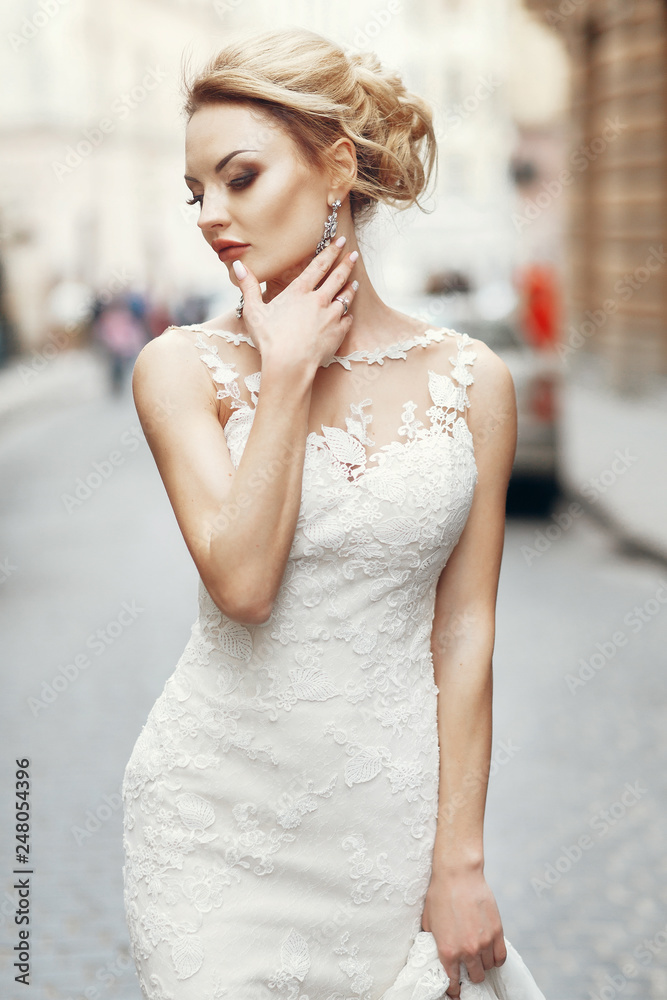 Fototapeta premium stylish bride posing on background of old wall in city street. luxury fashionable sexy woman in white dress walking in street