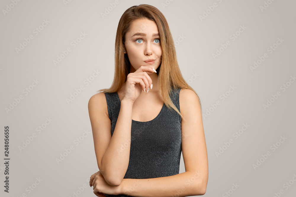 Photo of thoughtful young woman, holds hand to chin, considers something in mind, wears evening dress, stands against white background. People, thoughts, ideas Photo of thoughtful young woman, holds hand to chin, considers something in mind, wears evening dress, stands against white background. People, thoughts, ideas