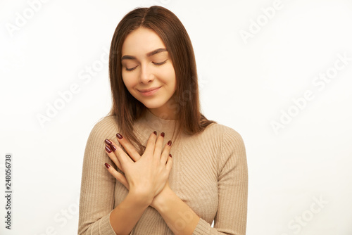 Pleased girl keeps hands together on chest, expresses gratitude, keeps eyes closed, touched with cordial congratulations, makes thankful gesture. Acknowledgment and appreciation concept 