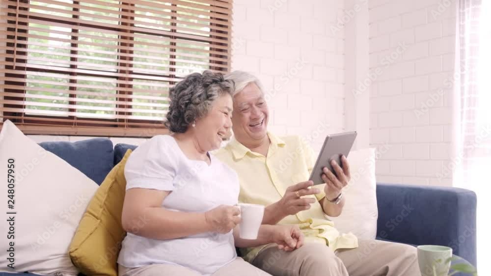 Asian elderly couple using tablet and drinking coffee in living room at home, couple enjoy love moment while lying on sofa when relaxed at home. Enjoying time lifestyle senior family at home concept. Asian elderly couple using tablet and drinking coffee in living room at home, couple enjoy love moment while lying on sofa when relaxed at home. Enjoying time lifestyle senior family at home concept.