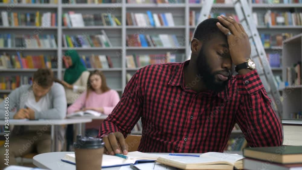 Overworked handsome african american student studying difficult university assignment, covering his forehead with arms, feeling fatigue and exhaustion during learning in academic library.