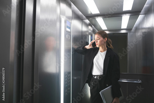 Dissatisfied businesswoman is talking on her smartphone in the elevator holding laptop in her hand. Frustrated office worker is having work problems and talking by phone keeping her notebook.