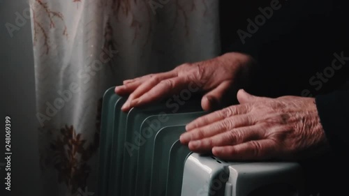 An elderly man warms his hands over an electric heater. In the off-season, central heating is delayed, so people have to buy additional heaters to keep houses warm despite increased electricity bills.