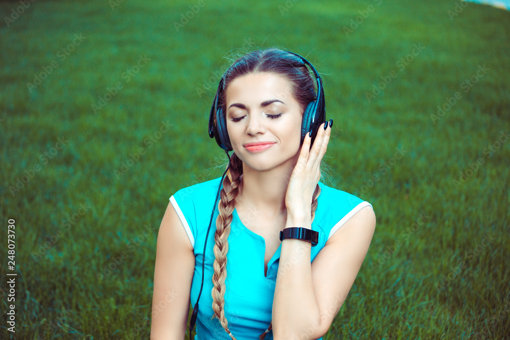 Shot of young fit sportswoman sitting on green grass meadow and listening to music enjoying it with eyes closed with headphones Shot of young fit sportswoman sitting on green grass meadow and listening to music enjoying it with eyes closed with headphones