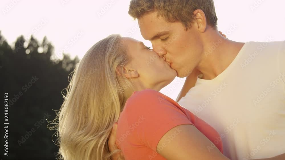 CLOSE UP, SLOW MOTION, SUN FLARE: Carefree young newlyweds enjoying an outdoor date and kissing in the romantic sunlit nature. Adorable shot of cheerful girl and boy kissing on a beautiful summer day. CLOSE UP, SLOW MOTION, SUN FLARE: Carefree young newlyweds enjoying an outdoor date and kissing in the romantic sunlit nature. Adorable shot of cheerful girl and boy kissing on a beautiful summer day.
