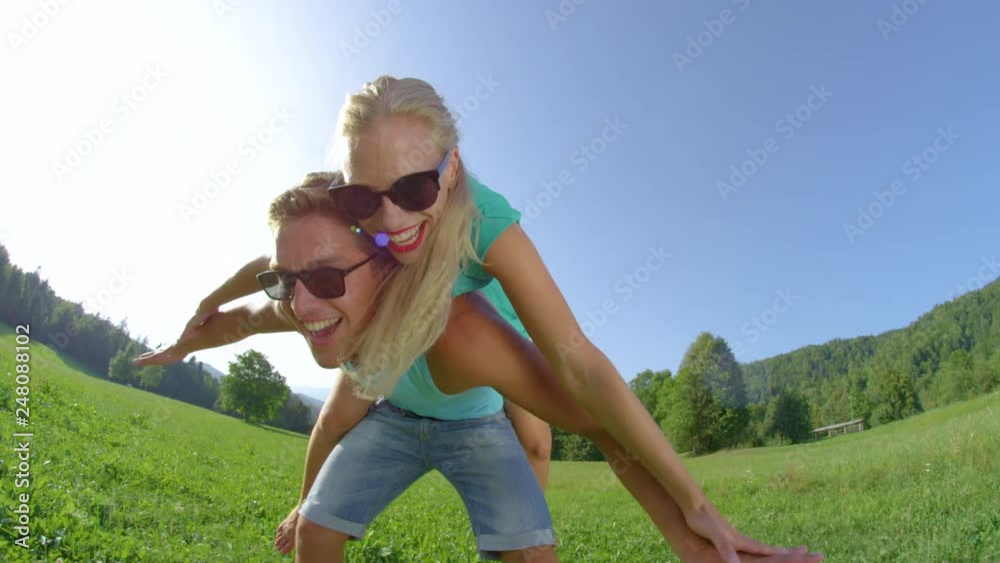 SLOW MOTION, SUN FLARE, PORTRAIT: Smiling young man gives his excited girlfriend a piggyback ride in sunny countryside. Handsome man sways while carrying the beautiful blonde haired woman on his back. SLOW MOTION, SUN FLARE, PORTRAIT: Smiling young man gives his excited girlfriend a piggyback ride in sunny countryside. Handsome man sways while carrying the beautiful blonde haired woman on his back.