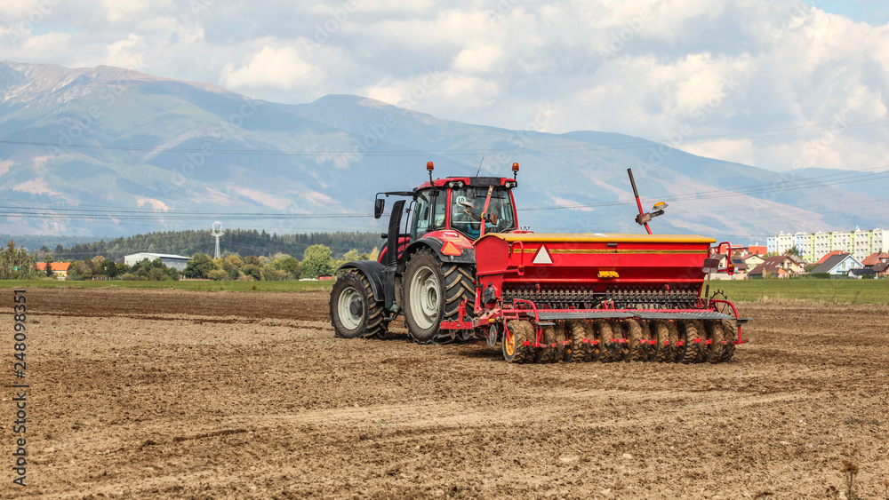 Fototapeta premium Red tractor pulling sowing trailer behind over empty field. Some village and mountains in background.