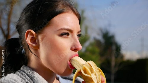 Slow motion, close-up attractive sexy young brunette woman with beautiful eyes and red lips eats banana on sunny day. Spring beriberi, vitamins in fruits.