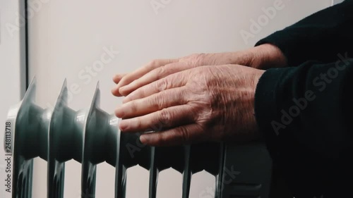 Senior elderly man warms his hands over electric heater. In the off-season, central heating is delayed, so people have to buy additional heaters to keep houses warm despite increased electricity bills