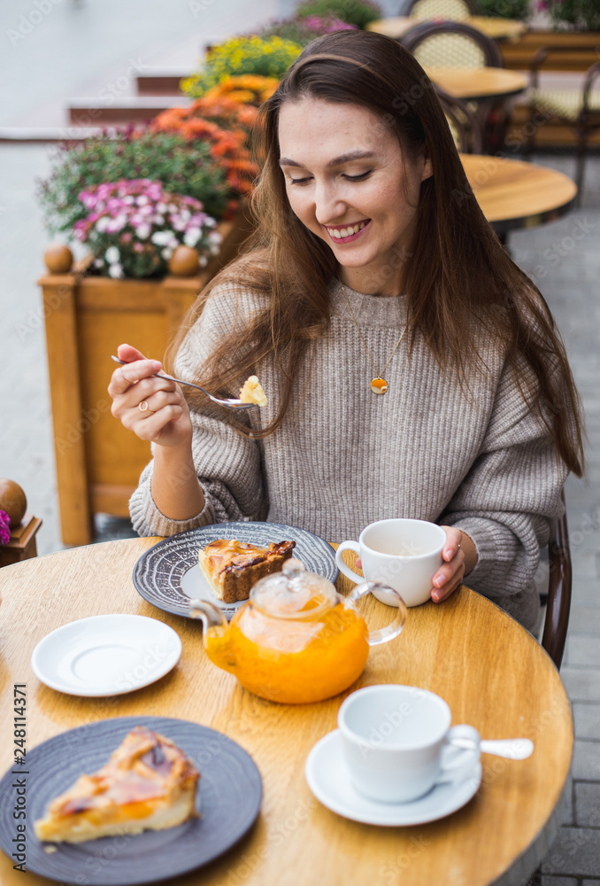 Young attractive smiling woman in gray knitted sweater drinking tea outdoors at the street cafe.