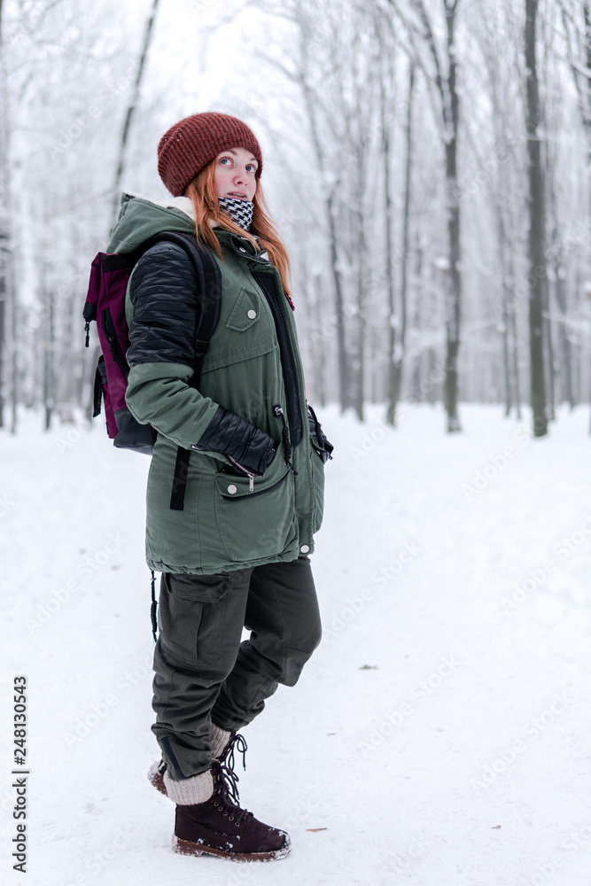 traveler girl with a bordo, red backpack walking on snow covered road in winter forest in frosty weather. vertical photo