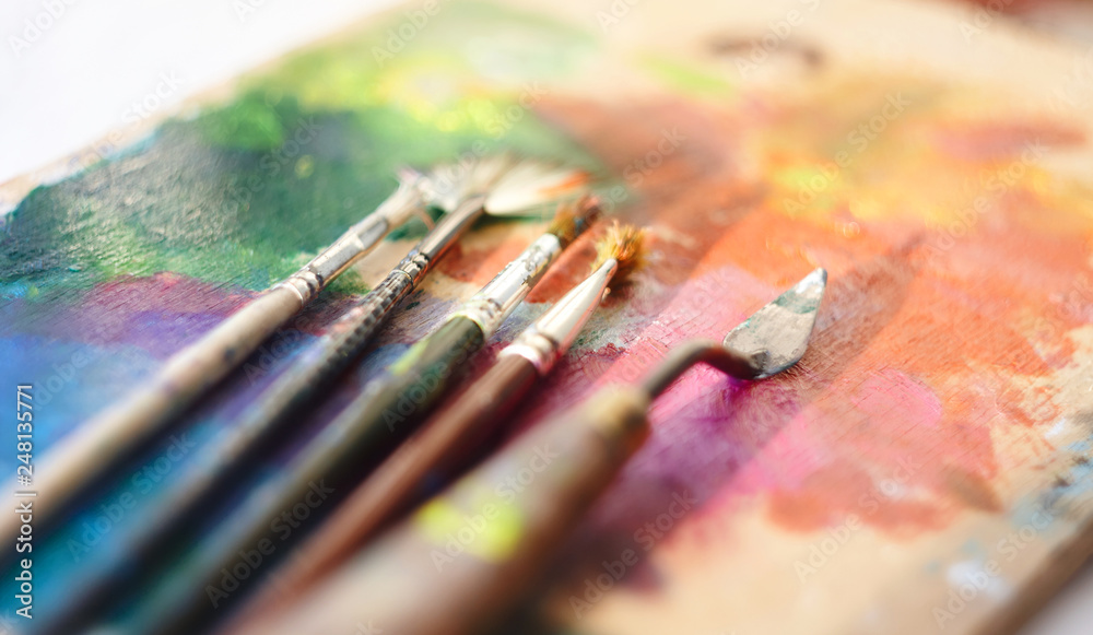 Brushes of different shapes and palette knife lie on a wooden palette, painted with oil paint of different colors of the rainbow