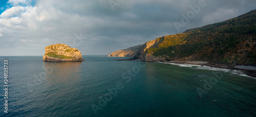 Aerial drone view of islet on the coast of Biscay with hermitage on the top; small church on the stone island, steep slope and rough coast in this area; craggy rocky uninhabited isle with old history photo