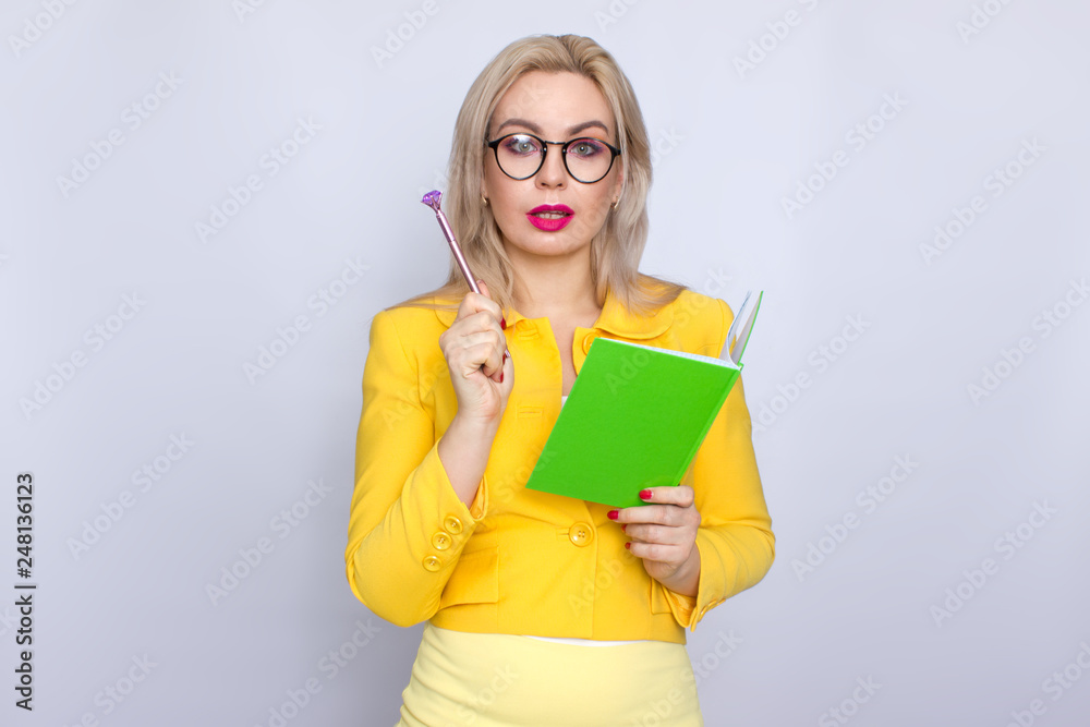 Close-up portrait of amazing blonde woman holding notebook, book, literature, in hands arms and pen pencil in yellow suit over white background in studio Close-up portrait of amazing blonde woman holding notebook, book, literature, in hands arms and pen pencil in yellow suit over white background in studio