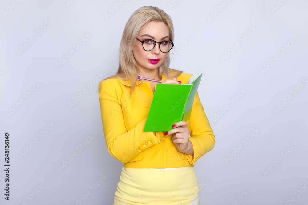 Close-up portrait of amazing blonde woman holding notebook, book, literature, in hands arms and pen pencil in yellow suit over white background in studio Close-up portrait of amazing blonde woman holding notebook, book, literature, in hands arms and pen pencil in yellow suit over white background in studio