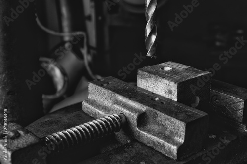 Black and white photo of drill bit coming down onto a piece of wood held in a vice in an industrial workshop photo