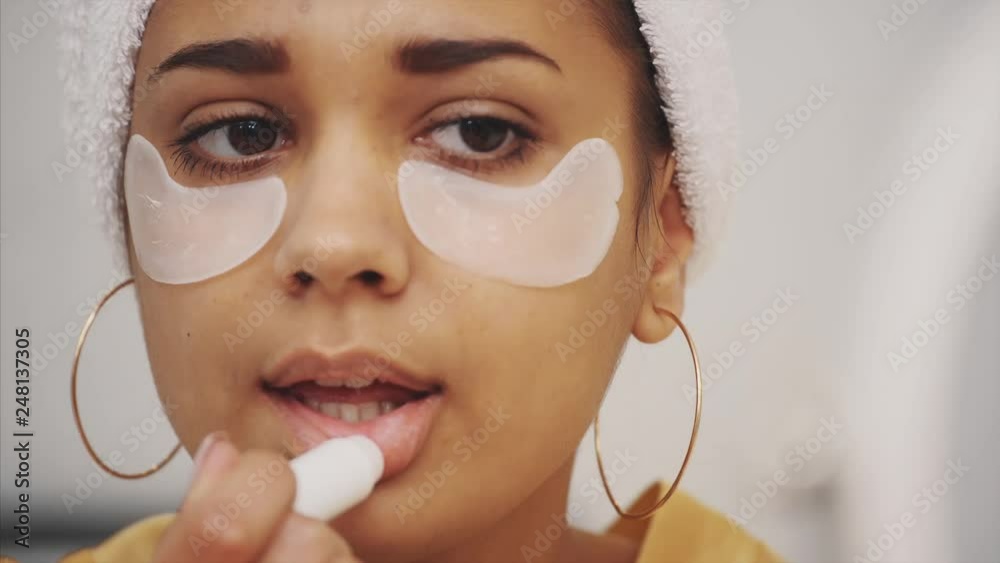 Closeup portrait of beautiful woman after bath with towel on head puts patches under the eyes from wrinkles and dark circles. Attractive female in bathrobe makes beauty treatments on the face at home.