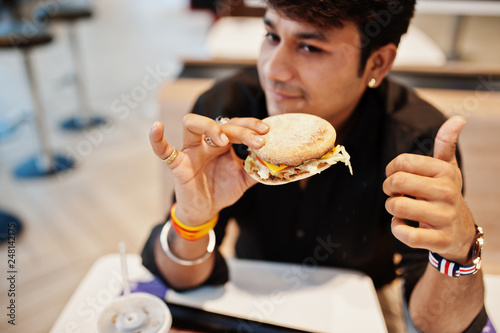 Stylish indian man sitting at fast food cafe and eating hamburger. Show thumb up.