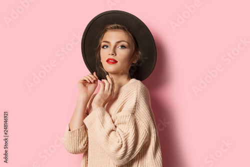 Portrait of glamour stylish model in beige cozy sweater and trendy hat. Attractive young woman posing on pink background. Modern fashion and autumn concept
