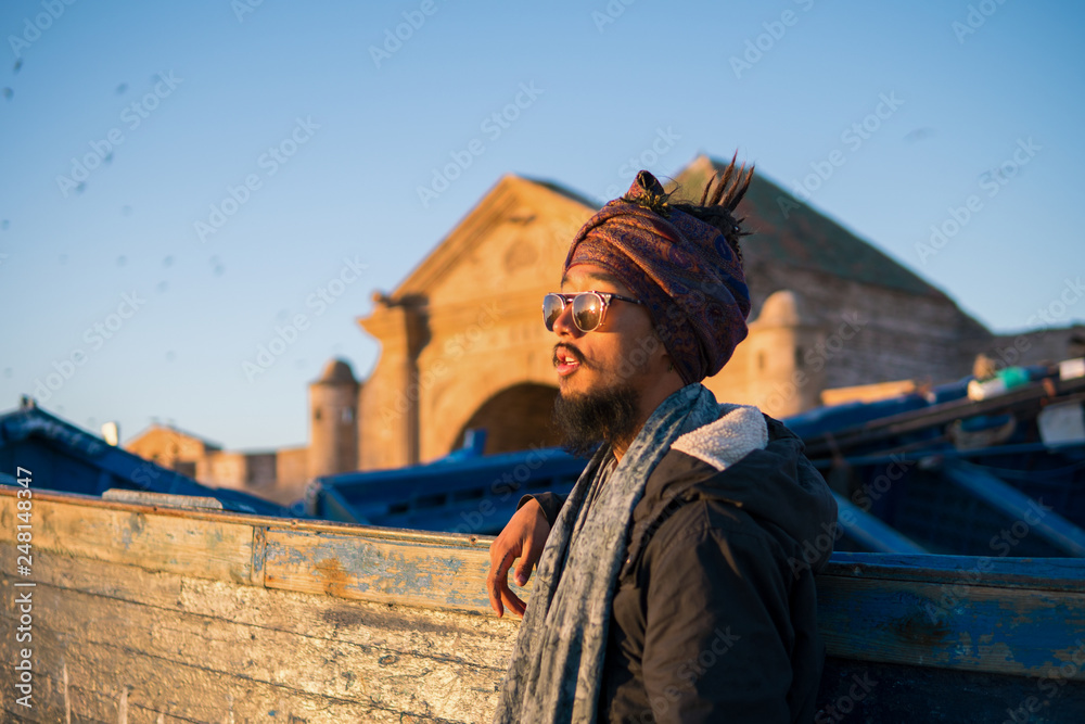 Young Asian Bearded Tourist man with Dreadlocks and scarf on head Stay hear the Blue Fisherman Boat in Essaouira, Morocco at the evening sunset time Young Asian Bearded Tourist man with Dreadlocks and scarf on head Stay hear the Blue Fisherman Boat in Essaouira, Morocco at the evening sunset time